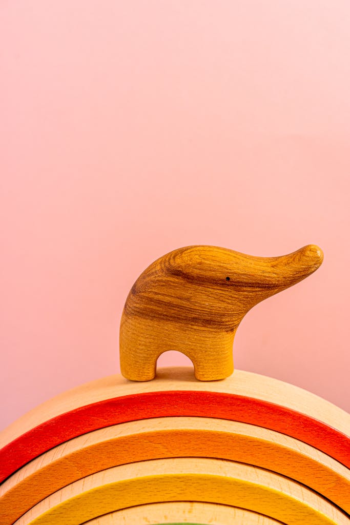 Charming wooden elephant toy on a colorful rainbow arch against a soft pink background.